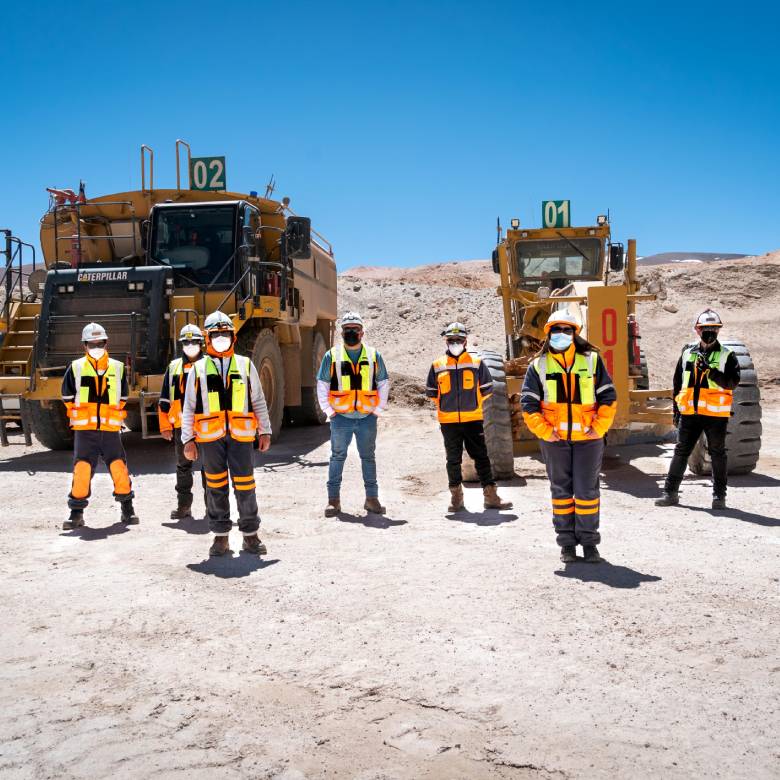 Seis trabajadores de minería con equipo de protección y máscaras en una mina a cielo abierto, de pie frente a dos grandes camiones Caterpillar amarillos numerados 01 y 02. El fondo es un paisaje desértico bajo un cielo azul, ilustrando un entorno de trabajo cubierto en los cursos online para aprender minería.