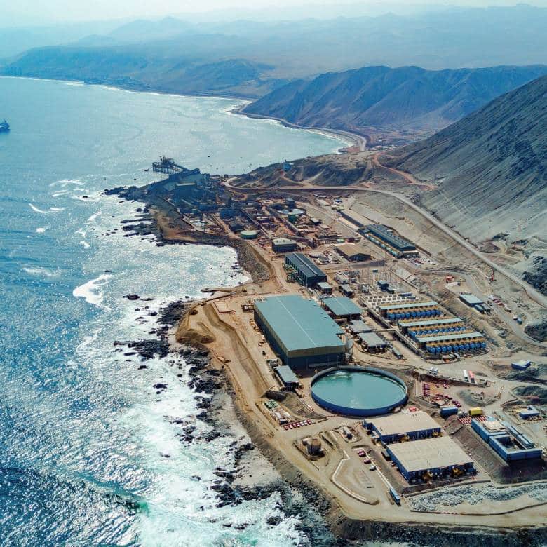 Fotografía aérea de un complejo industrial de minería costero con edificios operativos, un tanque circular y un muelle de carga situado entre montañas áridas y el océano.